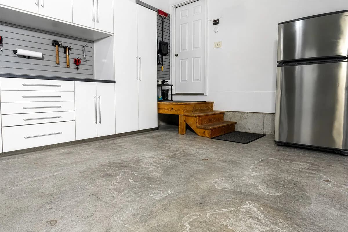Modern garage interior with gray concrete floor, storage cabinets, stairs, and a stainless steel fridge.